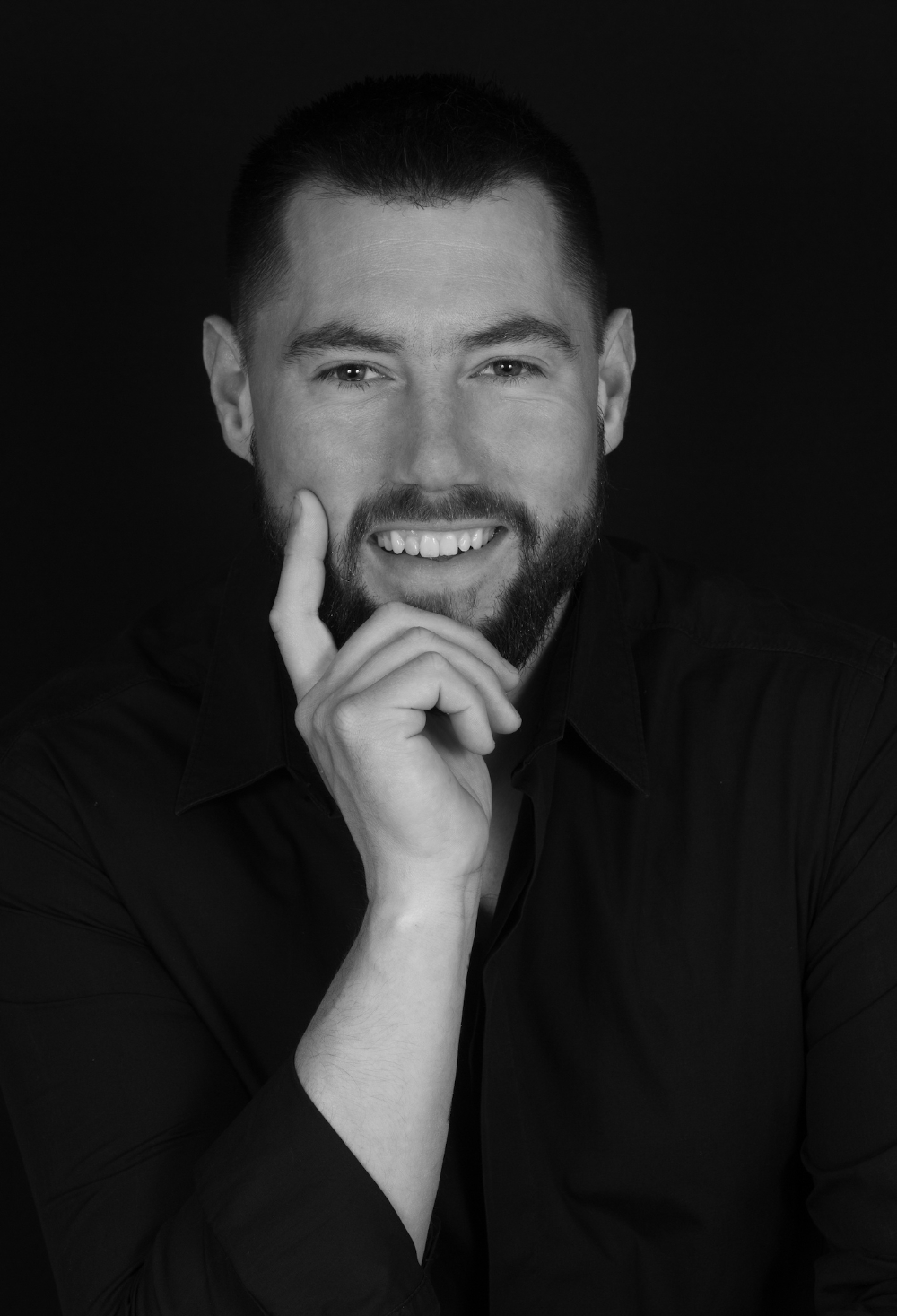 photo d'un jeune homme souriant en chemise noire sur un fond noir, une main au visage, l'index sur sa joue